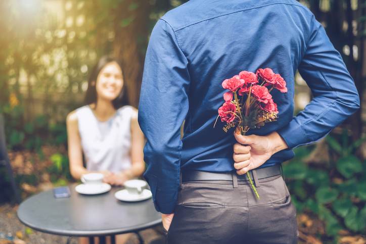 The man is hiding red flowers behind him in order to surprise his girlfriend. The man is hiding red flowers behind him in order to surprise his girlfriend.
